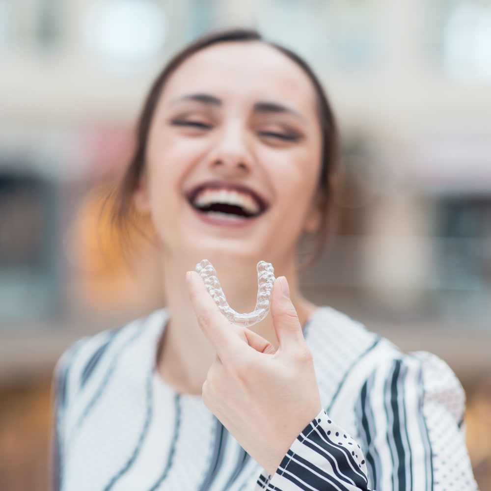 A woman stands just out of focus smilng while holding up her clear aligner which is in focus.
