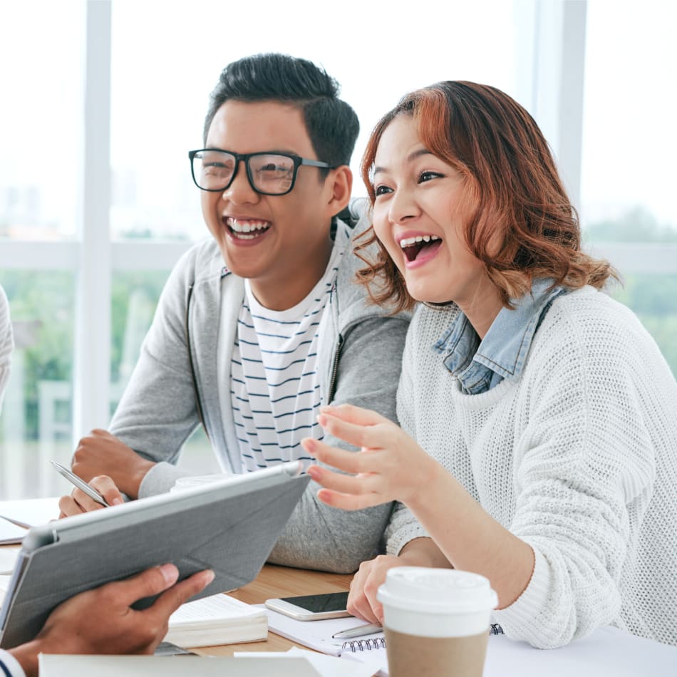 A man and a woman sit inside laughing and looking at a tablet being held by someone off camera.