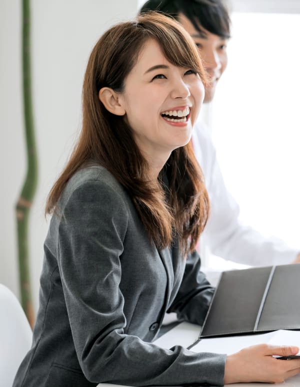 Young woman sits facing the right and smiling while a man can be seen just behind her.