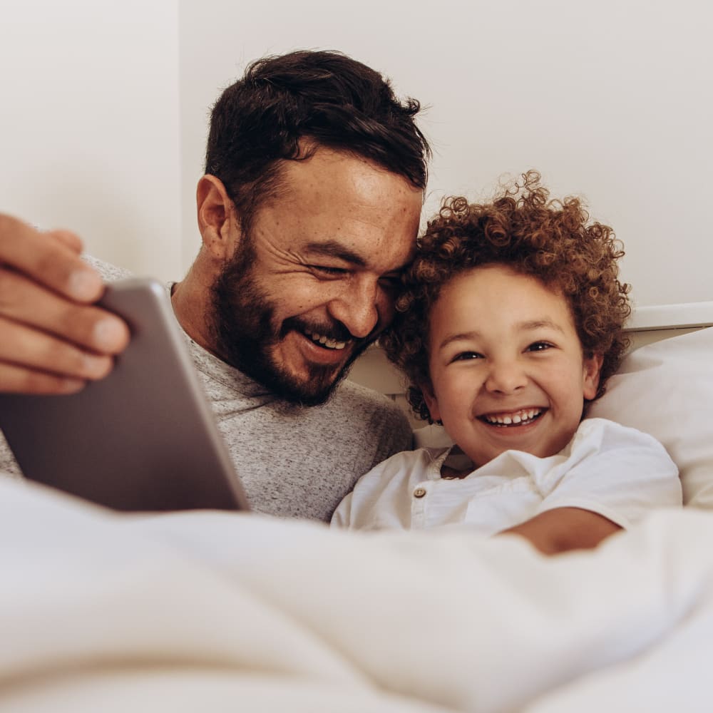 A man and a small boy lay in bed smiling and laughing while looking at a tablet.