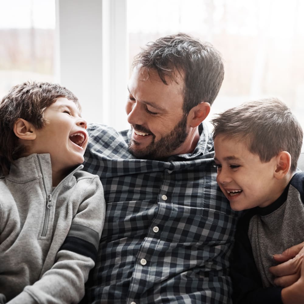 A man and two young boys sit on a sofa, looking at eachother and laughing.