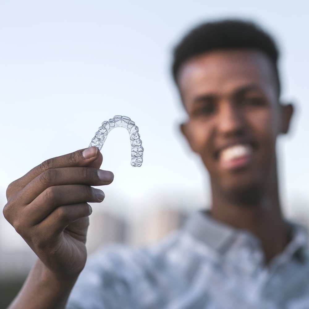 A teen boy stands just out of focus while holding his clear aligner in focus up in front of him.